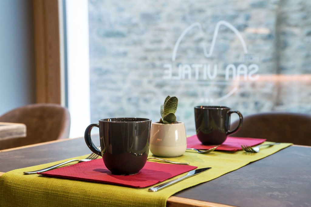 A dining table with two dark mugs, a small potted plant, and a red placemat.