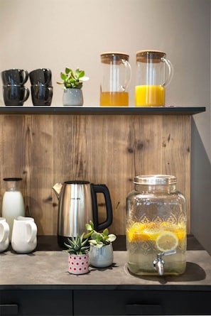 A kitchen counter with a coffee maker, a glass jar with a lemon slice, and a shelf with mugs and a small plant. - Hotel San Vitale (Bormio)