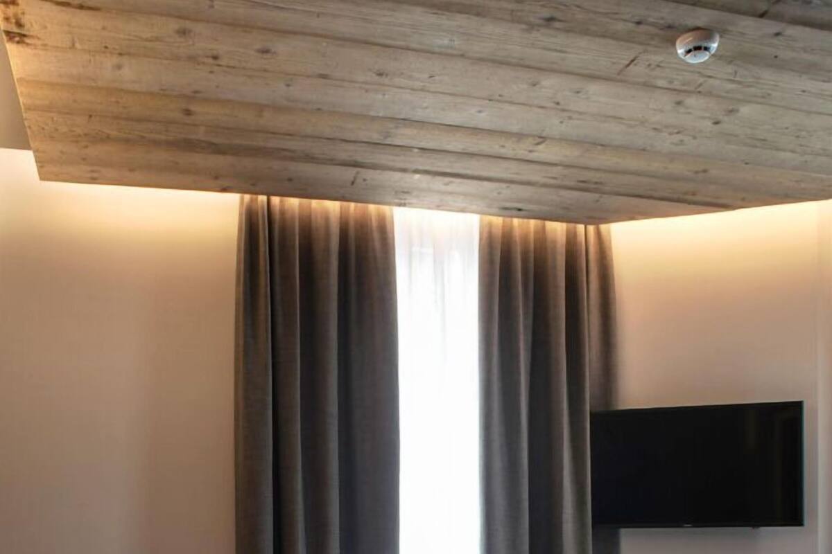 A bedroom with a bed, curtains, a mounted TV, and a wooden ceiling.