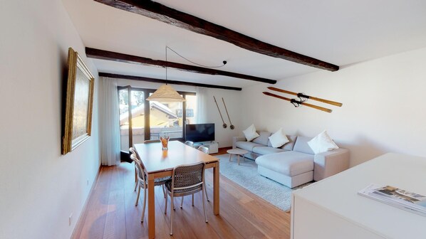 Ein modernes Wohnzimmer mit Essbereich, weißen Möbeln, Holzbalken und Skiausrüstung an der Wand.