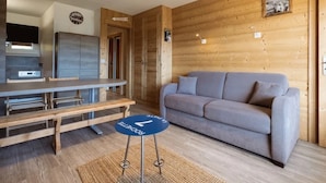 A modern living room with a grey sofa, wooden walls, a kitchen area with a fridge and cabinets, and a small round table with a blue coaster. - 3* Apartment, sleeps 6, pets allowed, garage, mountain view (La Clusaz)