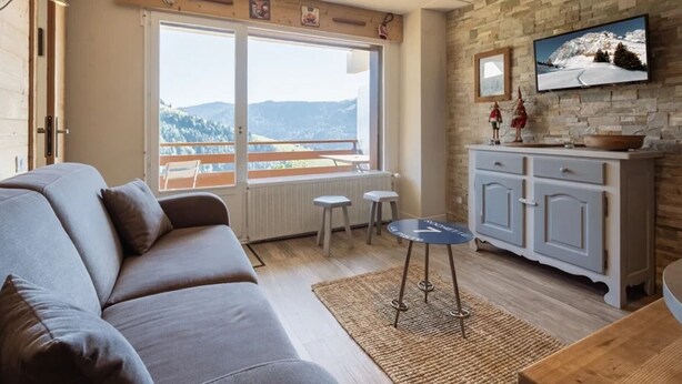 A modern living room with a sofa, a small table, and a view of mountains through large windows. - 3* Apartment, sleeps 6, pets allowed, garage, mountain view (La Clusaz)