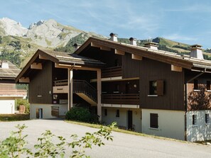 A two-story wooden building with a balcony, a staircase, and a mountain backdrop. - 3* Apartment, sleeps 6, pets allowed, garage, mountain view (La Clusaz)