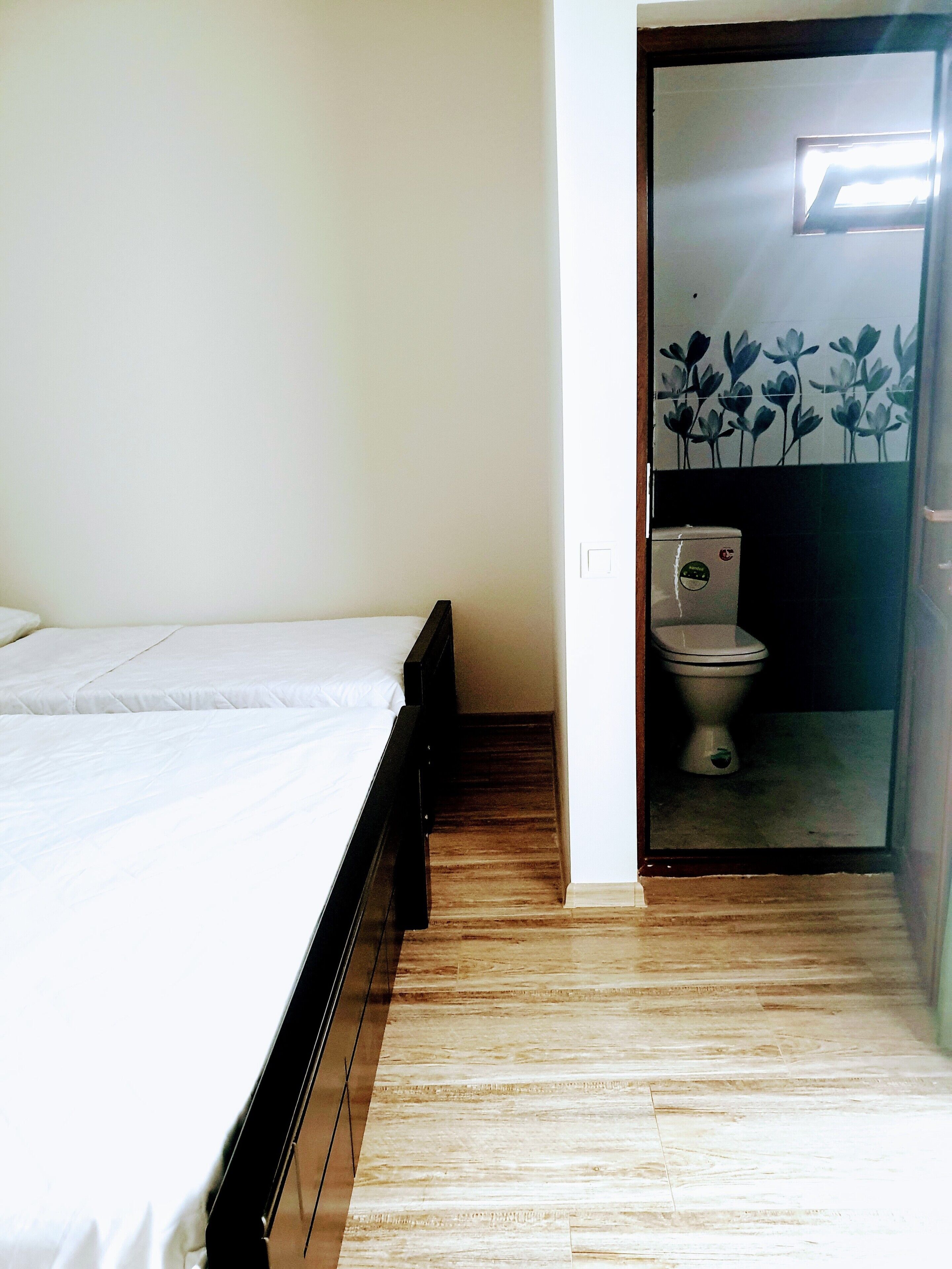 A bedroom with a wooden floor, a bed with white bedding, and a view of a bathroom through an open door.