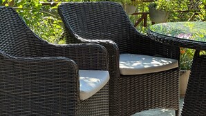 Wicker chairs with cushions around a table in an outdoor garden area with lush greenery and flowering plants.