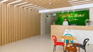 Zona de recepció d'hotel amb sostre de lamel·les de fusta, taulell de recepció amb paret verda i el rètol 'Prime Hotel', i diverses cadires i taules.