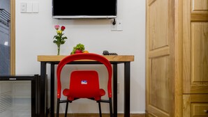 Habitació senzilla amb escriptori de fusta, una cadira vermella, taula petita amb un ram de flors, televisió penjada a la paret i armari de fusta.