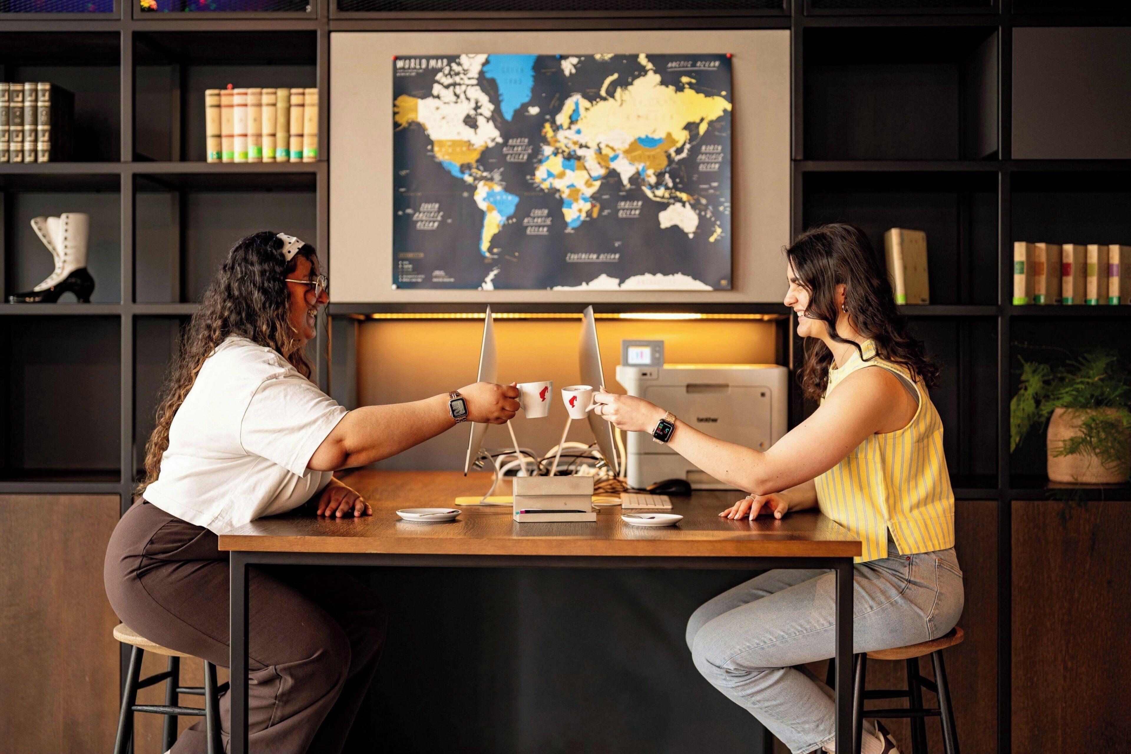 Two individuals seated at a wooden table with a world map on the wall behind them.