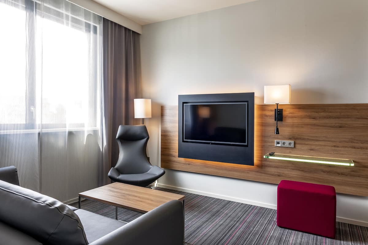 Una habitación de hotel moderna con un televisor de pantalla plana grande montado en la pared, un sofá cómodo, una mesita pequeña y un otoman rojo.