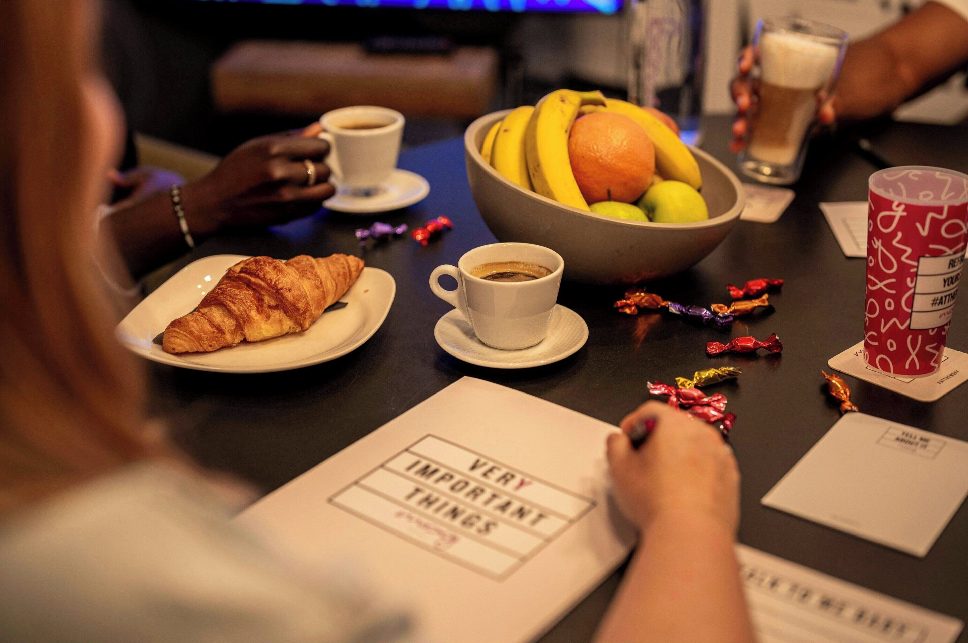 Un tavolo da pranzo con un croissant, una tazza di caffè, una ciotola di frutta e un bicchiere di bevanda.