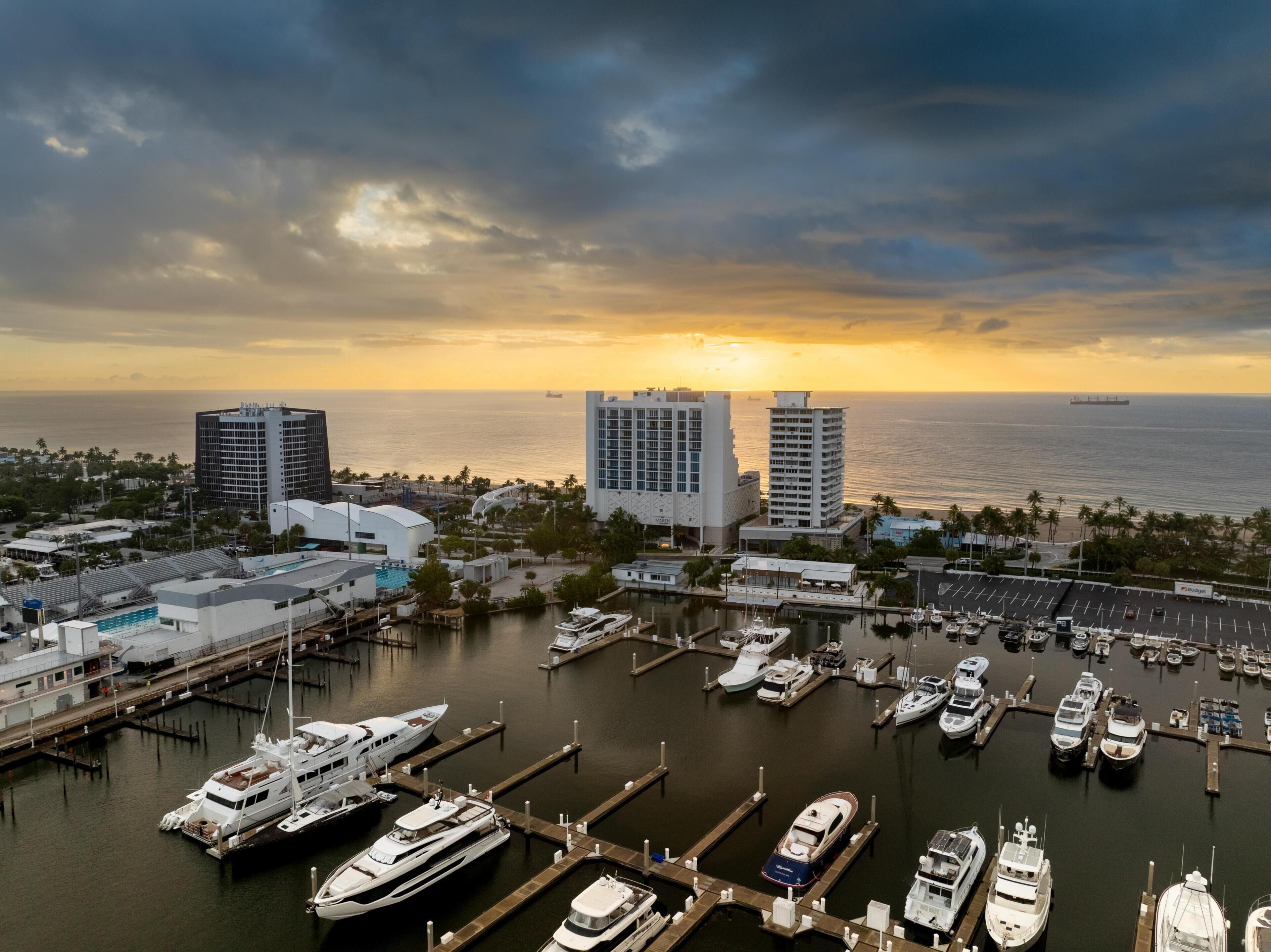 A marina with multiple boats docked, a large building with a curved roof, and a sunset over the ocean.