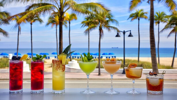 A row of cocktails on a table with a beach and palm trees in the background.