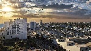 A coastal cityscape with a prominent hotel featuring a modern design, a marina with numerous boats, and a beach with palm trees.