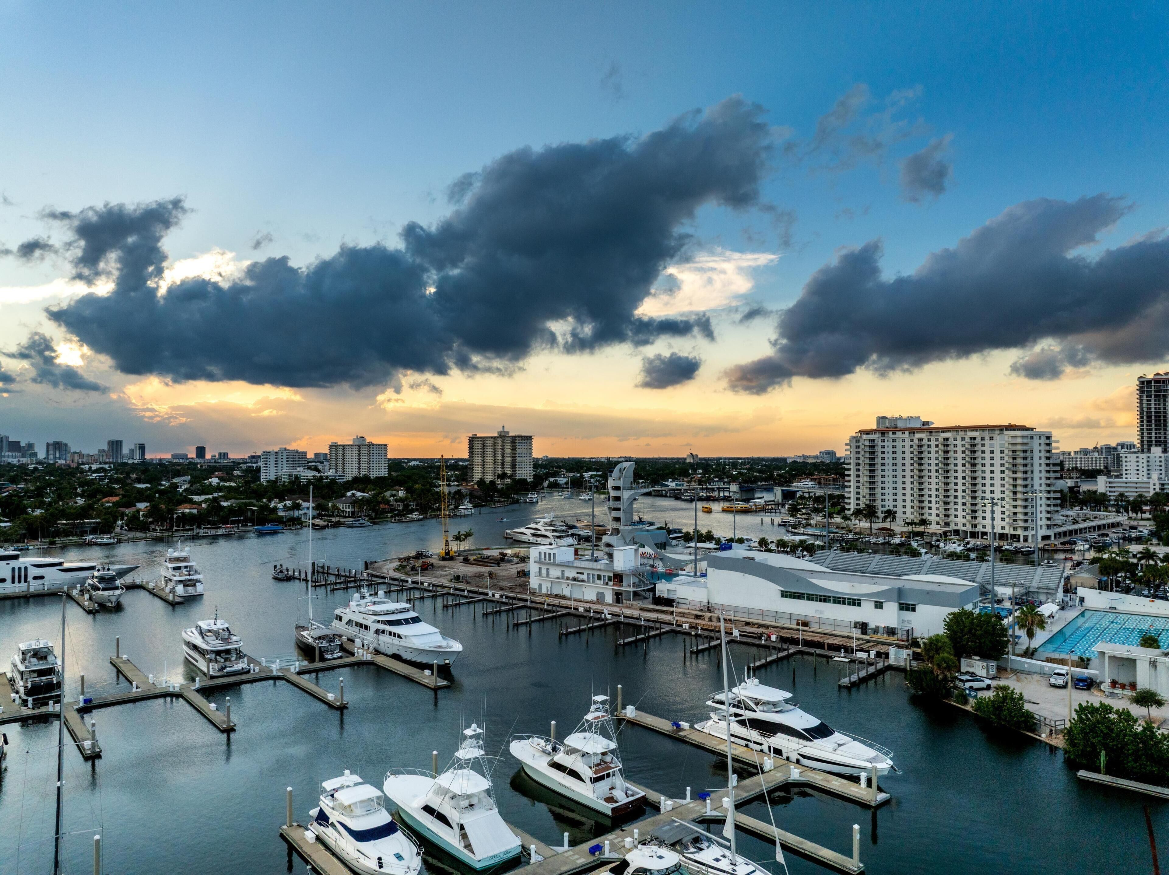 A marina with multiple boats docked, a large building with a flat roof, and a city skyline in the background.