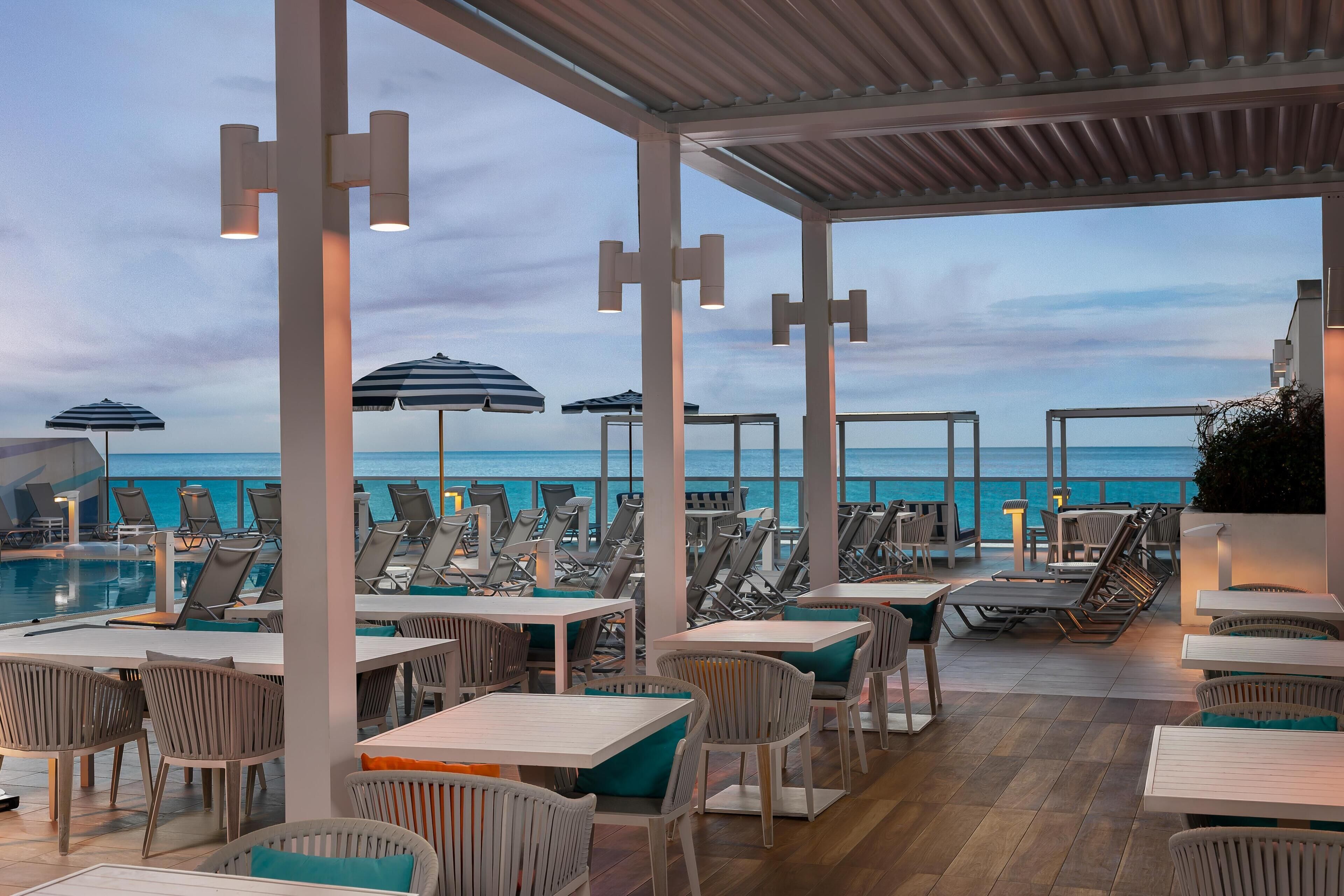 A poolside area with lounge chairs and tables, overlooking the sea.