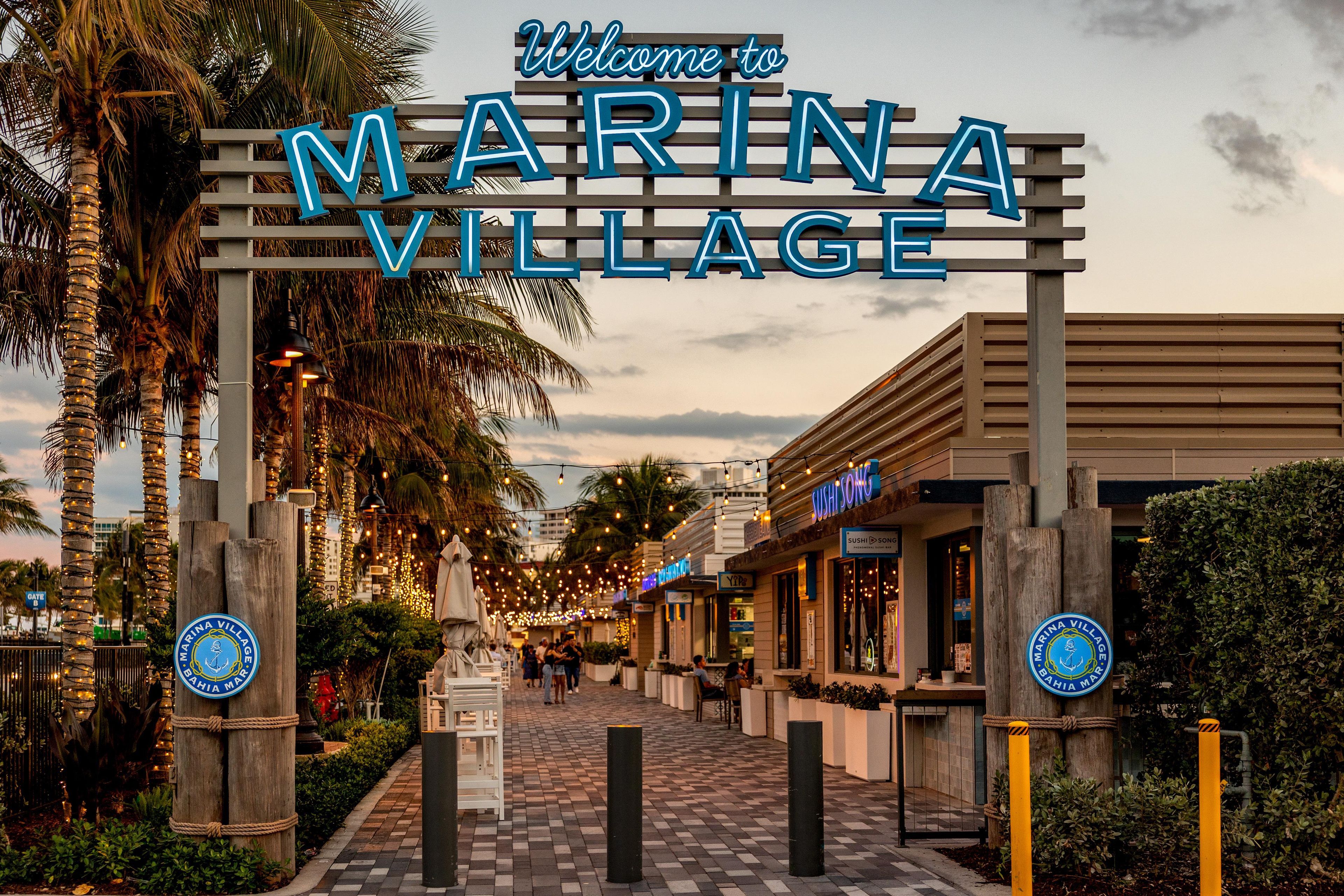 Un letrero da la bienvenida a Marina Village, con un arco de madera, palmeras y un camino pavimentado.