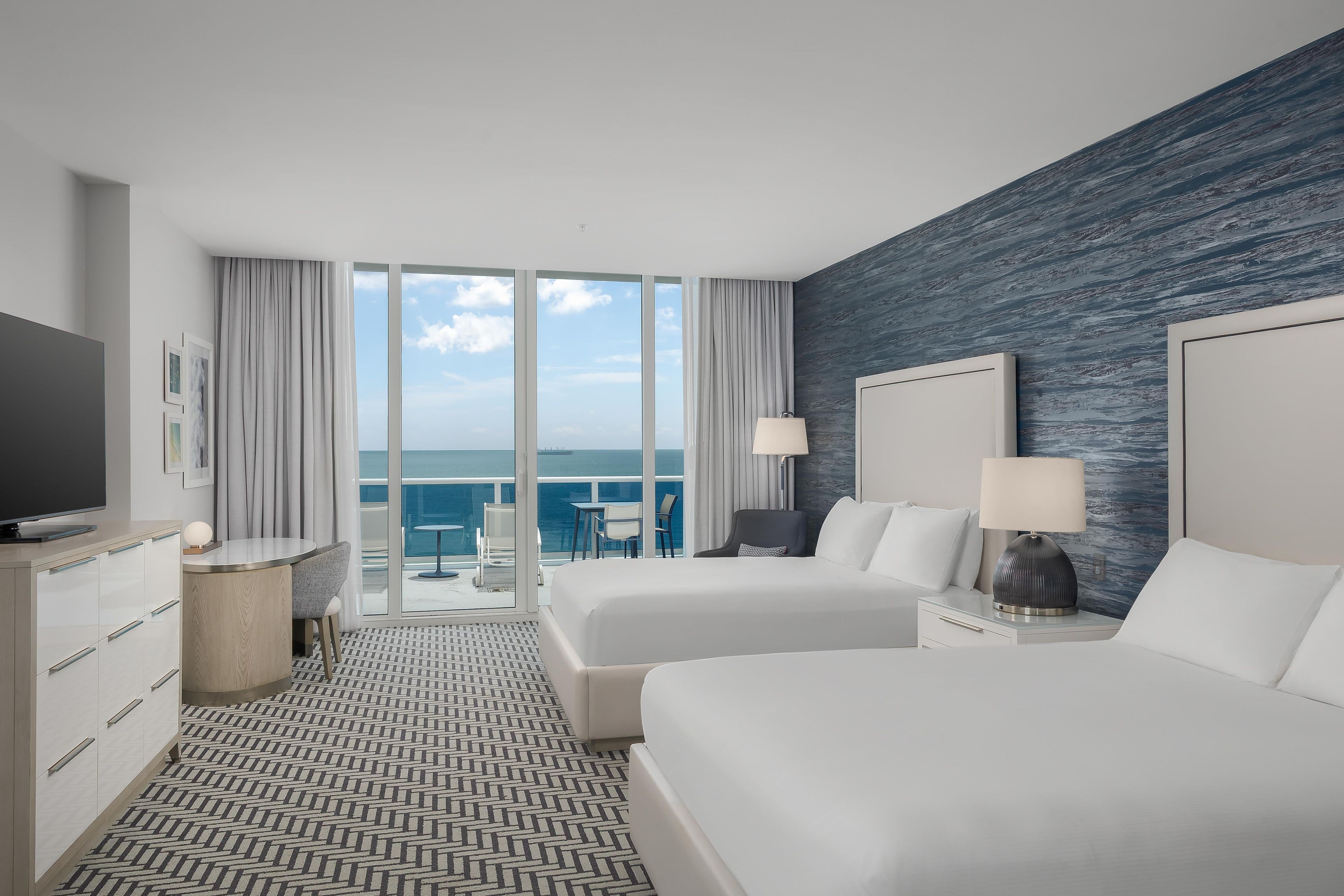 A hotel room with two beds, a TV, a desk, and a view of the ocean.