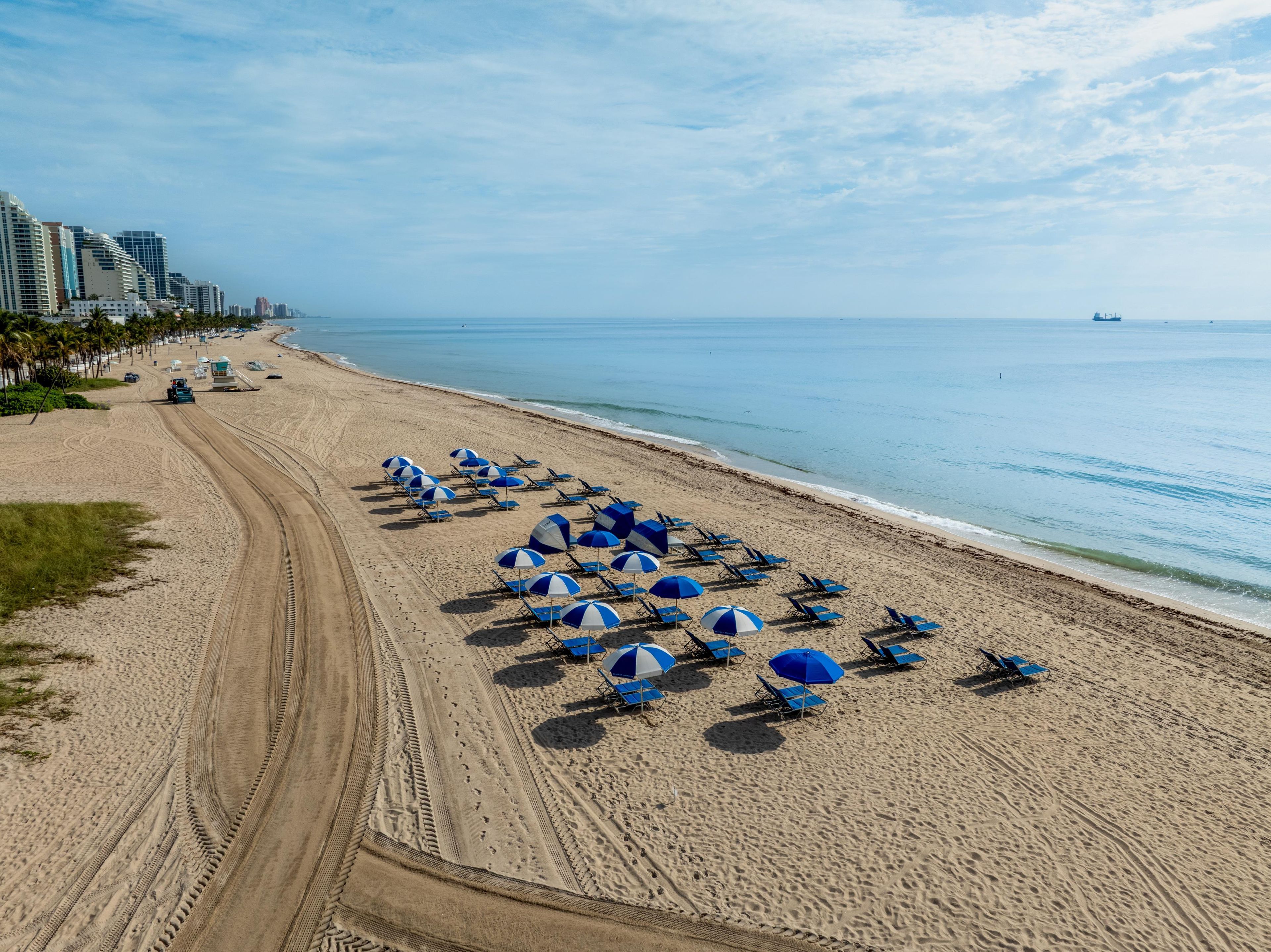 Une plage bordée de rangées de chaises de plage et de parasols bleus, un sentier de terre, et des immeubles de grande hauteur en arrière-plan.
