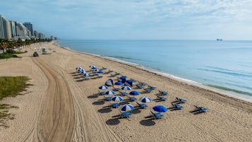 Une plage bordée de rangées de chaises de plage et de parasols bleus, un sentier de terre, et des immeubles de grande hauteur en arrière-plan.