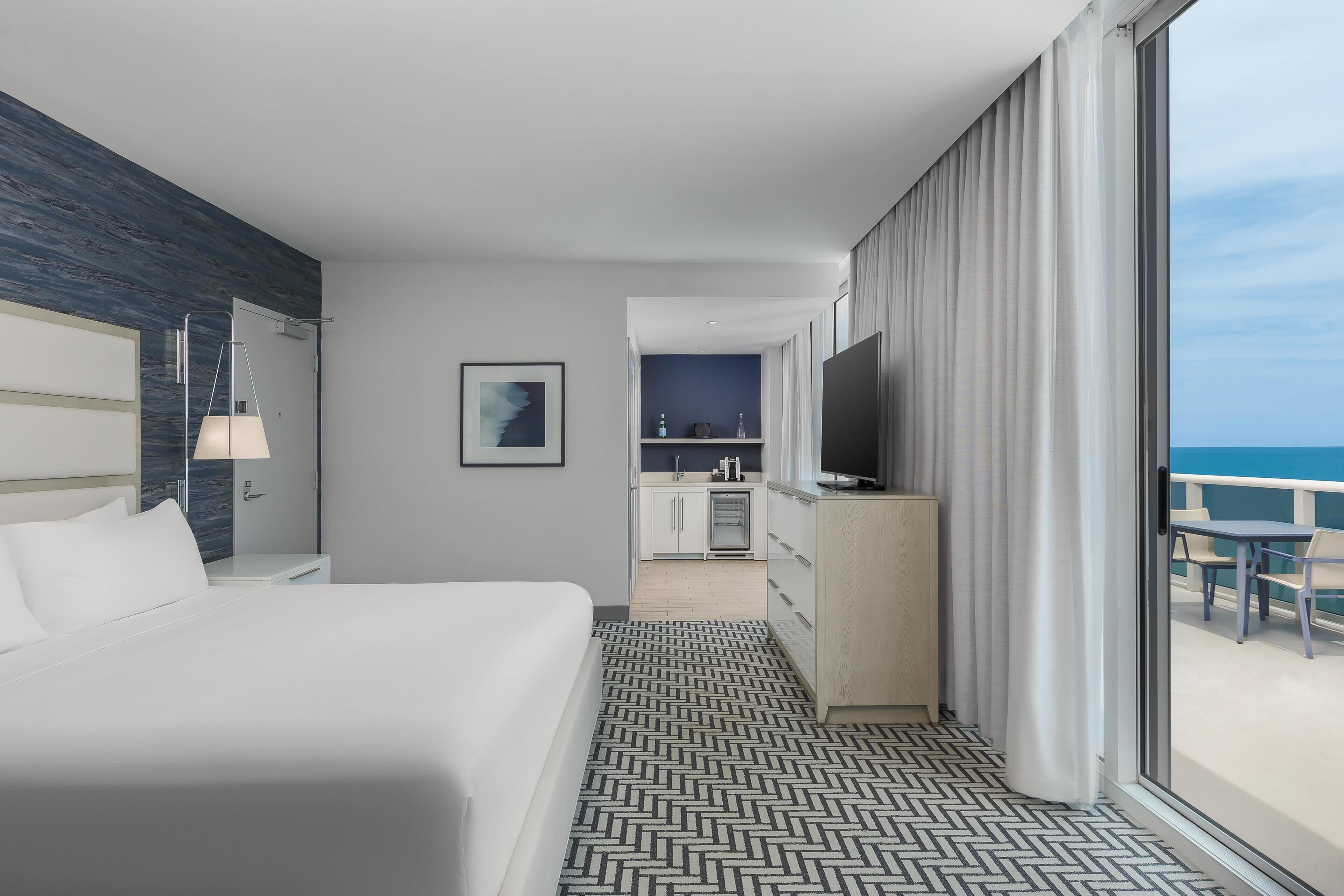 Ein Hotelzimmer mit Bett, Schreibtisch, Fernseher, Meerblick und einer Küchenzeile.