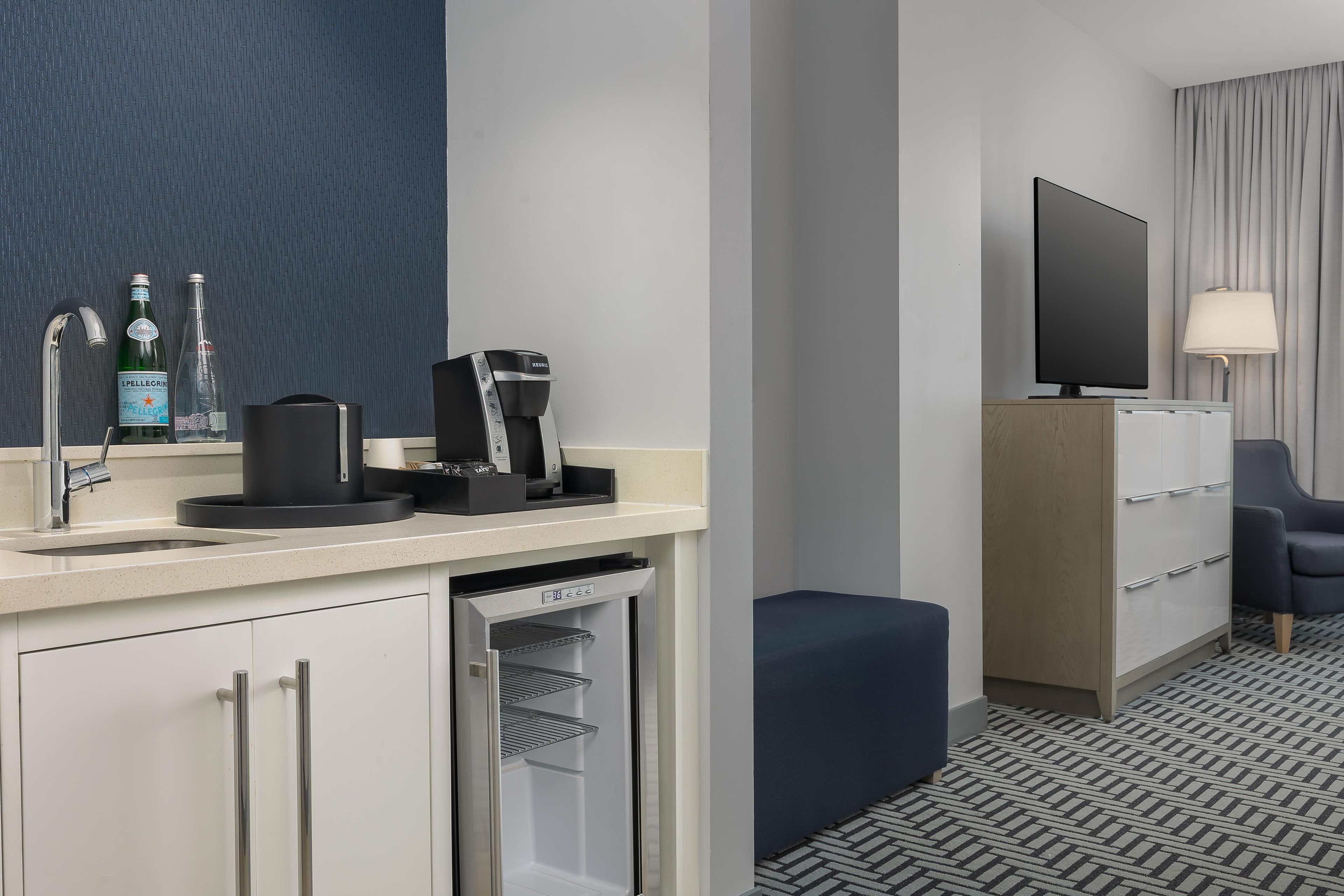 A hotel room with a kitchenette, a flat-screen TV, a sofa, and a patterned floor.