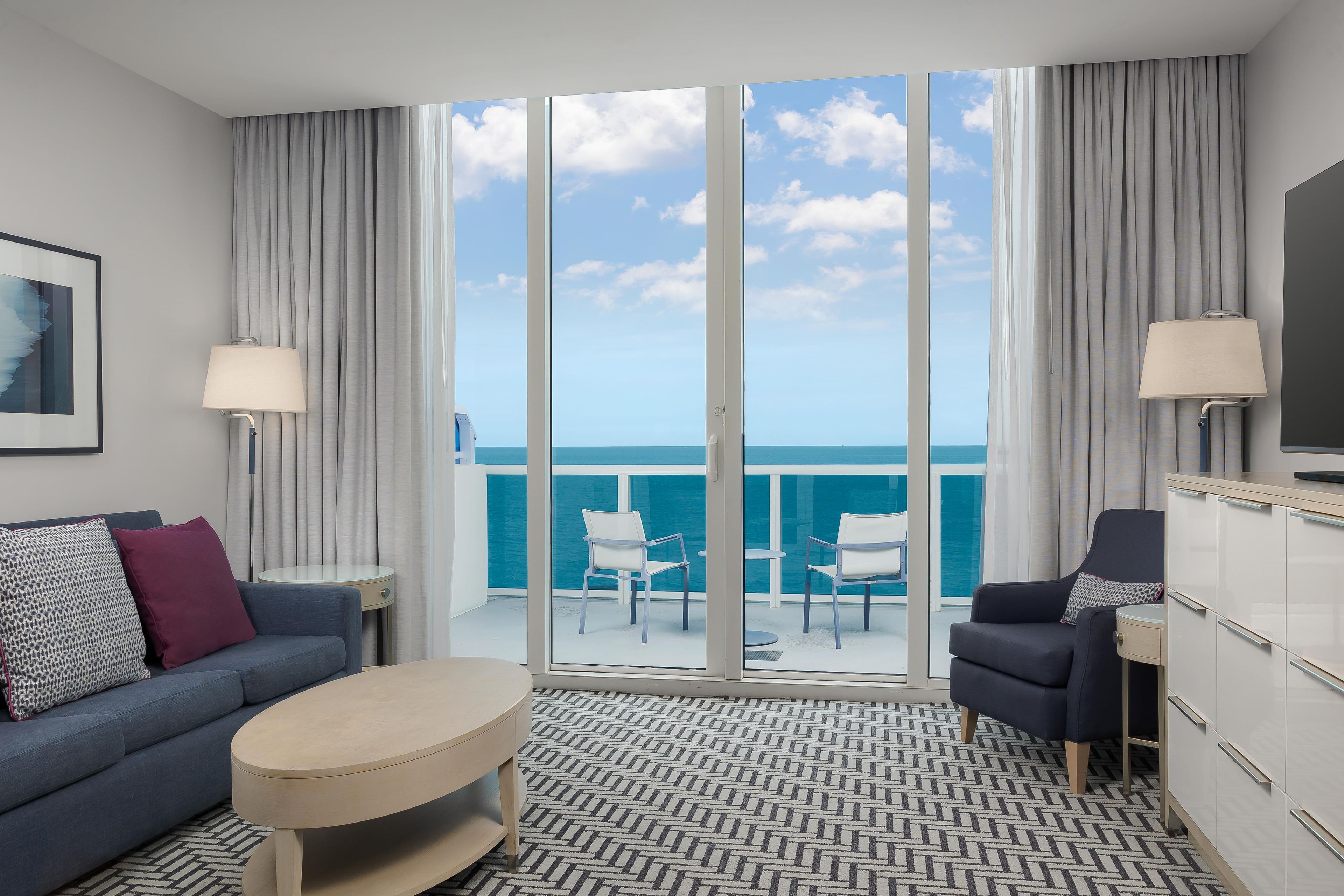 A modern hotel room with a balcony, a sofa, a coffee table, a TV, and a view of the ocean.