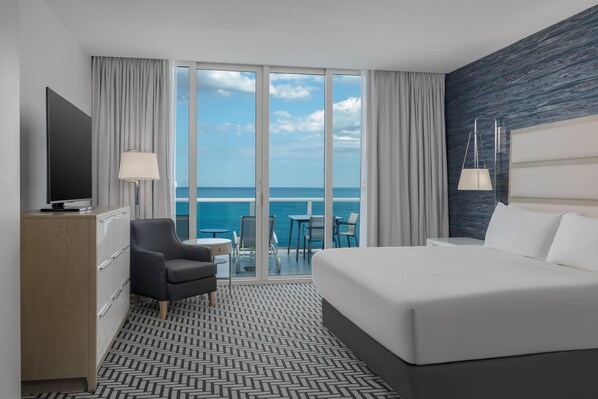 A hotel room with a large bed, a desk, a chair, a TV, and a view of the ocean.