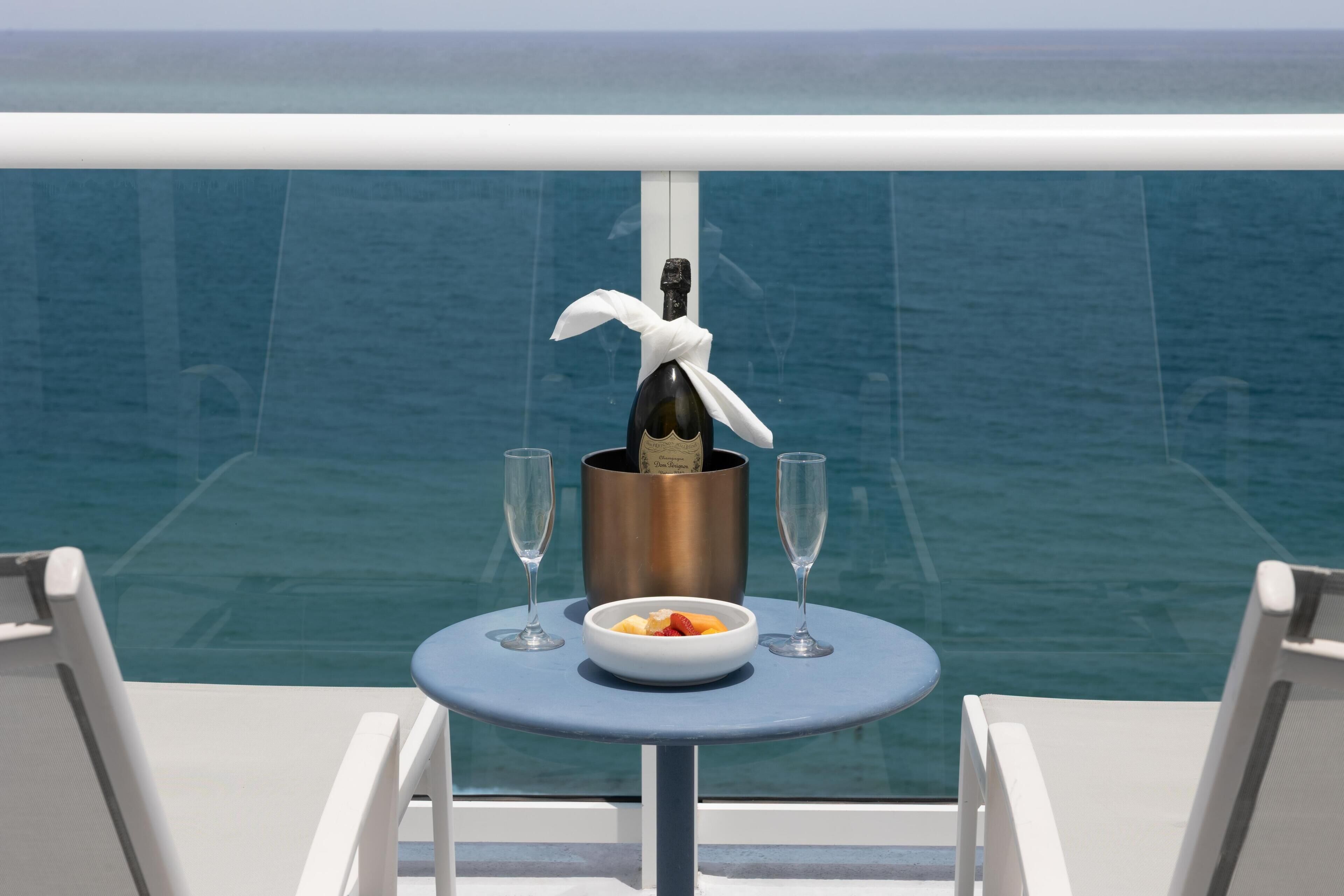 A table with a bottle of champagne, two glasses, and a bowl of fruit on a balcony with a view of the ocean.