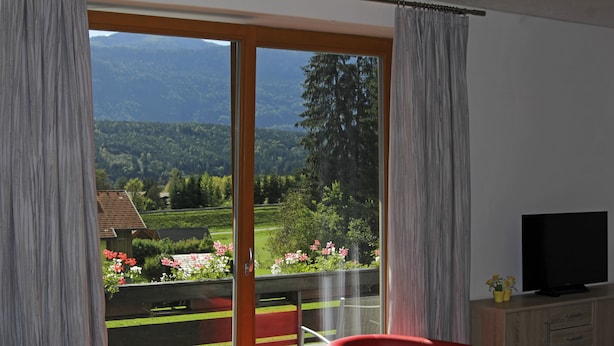 Una habitación con balcón, una silla roja, un televisor sobre un mueble de madera y vistas a un bosque y montañas.
