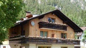 Un chalé de madera con balcón, ventanas amplias y un patio con mobiliario exterior.
