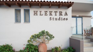The exterior of HLEKTRA Studios, featuring a white facade, two blue-framed windows, a large terracotta pot with a cactus, and a staircase leading to a glass door. - Electra Studios (Platanias)