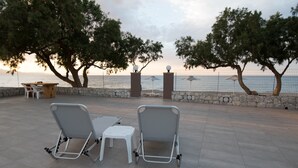 An outdoor seating area with two metal chairs and a small table, overlooking a body of water with a stone wall and trees. - Electra Studios (Platanias)