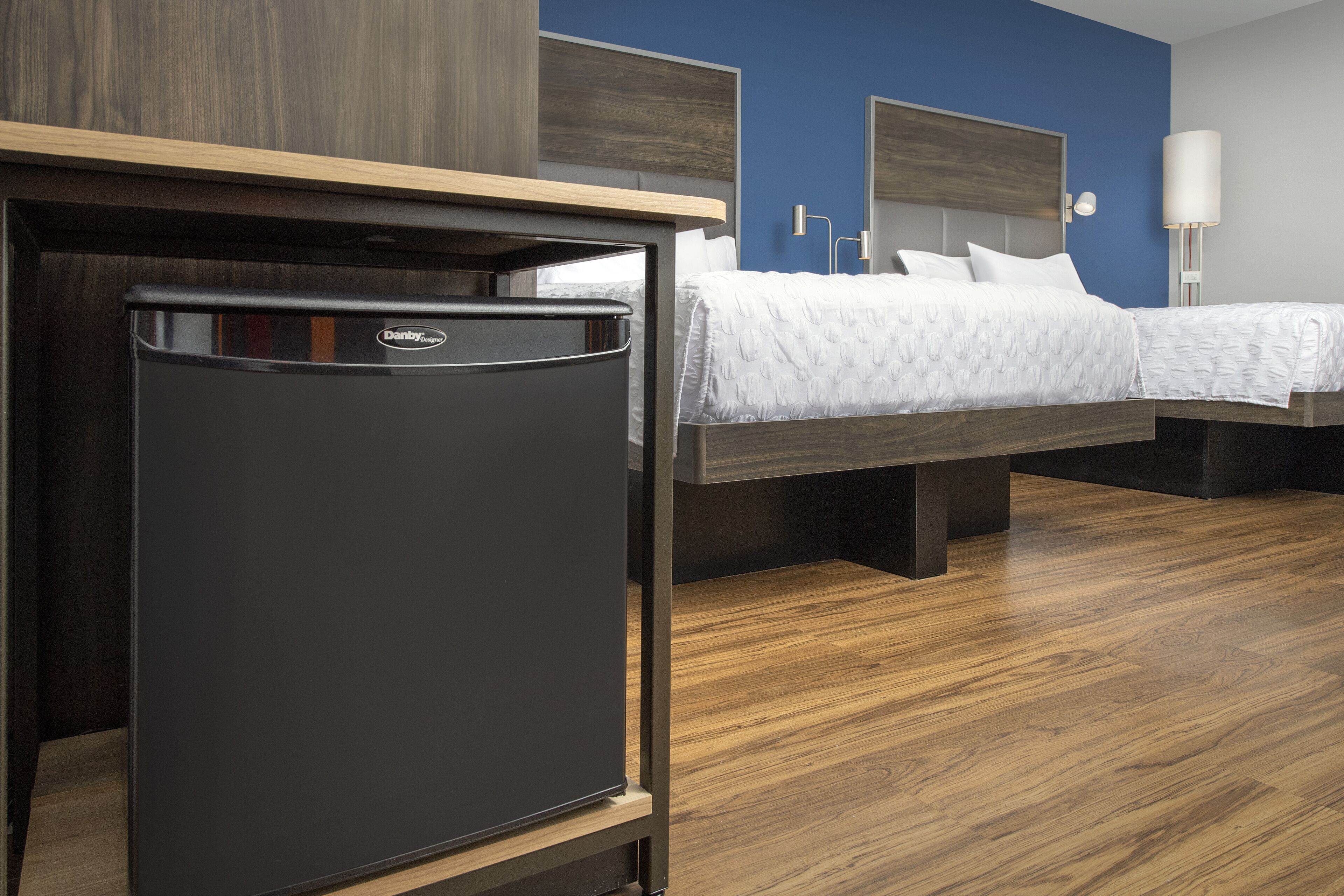 Une chambre d’hôtel moderne équipée d’un petit réfrigérateur, d’un lit avec une literie blanche et d’un plancher en bois.