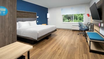 A modern hotel room with a bed, a desk, a chair, a TV, and a window with a view of greenery.