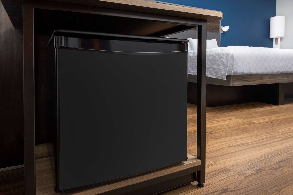 Een kleine, donkere koelkast in een moderne hotelkamer met houten vloeren en een bed met witte beddengoed.