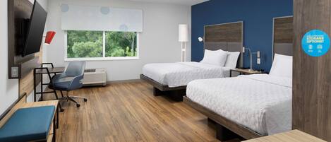 A hotel room with two beds, a desk, a chair, a TV, and a window with a view of greenery.