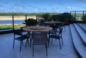 A terrace with round tables and chairs, overlooking a body of water and greenery. - FlatBeachBarra - Love Island - Paiva Beach (Barra de Jangada)