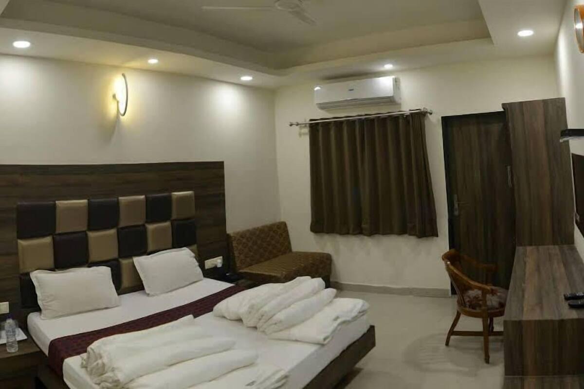 A hotel room with a large bed, a wooden headboard, a desk, a chair, a nightstand, an air conditioner, and a wall-mounted light fixture.
