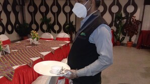 A person in a professional uniform serving at a table with a red tablecloth and white napkins, in a room with patterned walls and potted plants. - Hotel Shree Vatika (Bhopal)