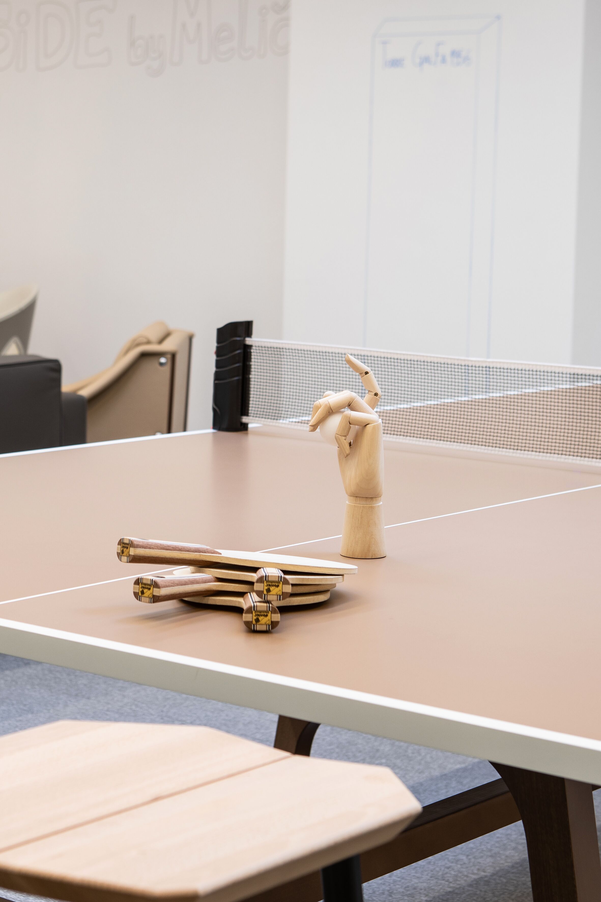 A ping pong table with a wooden hand model and paddles in the center, a whiteboard in the background, and a black chair to the left.