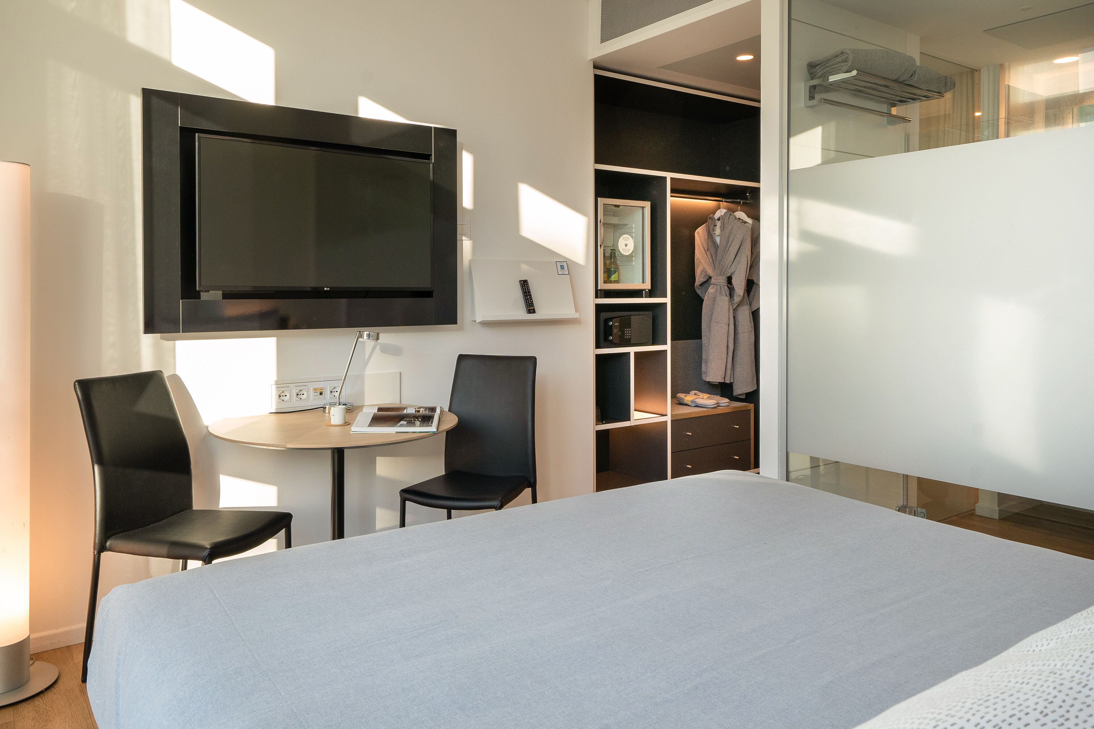 Quarto de hotel moderno com uma cama grande, uma TV fixada na parede, uma escrivaninha com cadeira e um closet espaçoso.