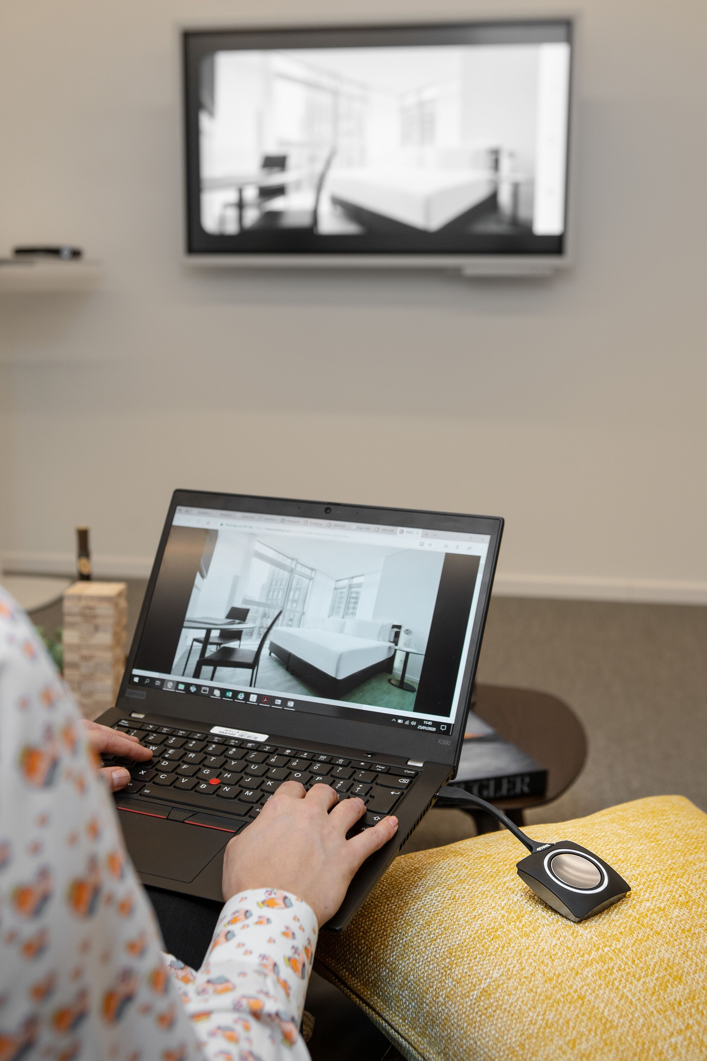 Uma pessoa usando um laptop em um sofá amarelo, com uma pequena mesa ao lado. Uma televisão está montada na parede ao fundo.