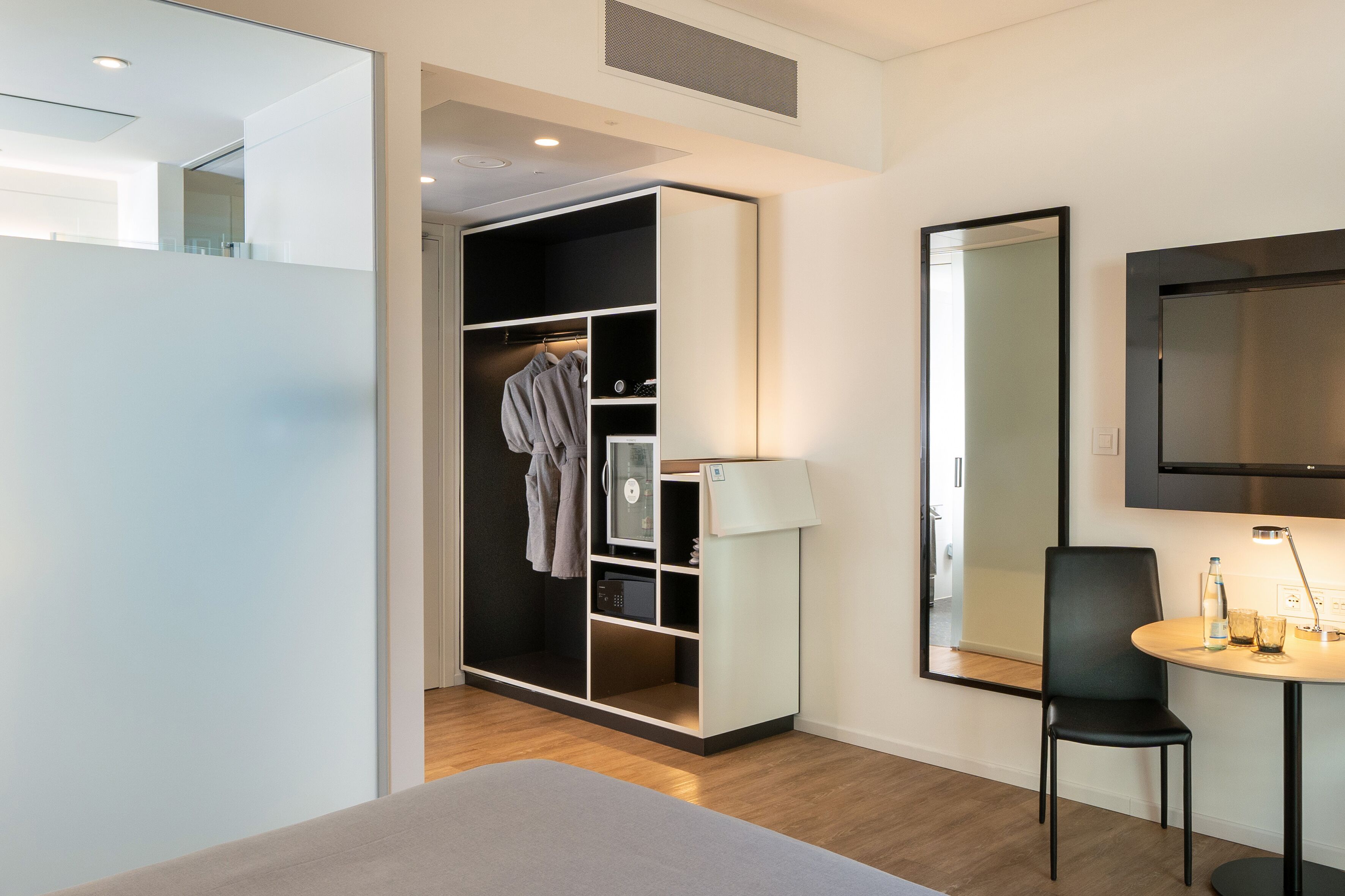 A modern hotel room with a large bed, a walk-in closet, a desk with a chair, and a TV mounted on the wall.