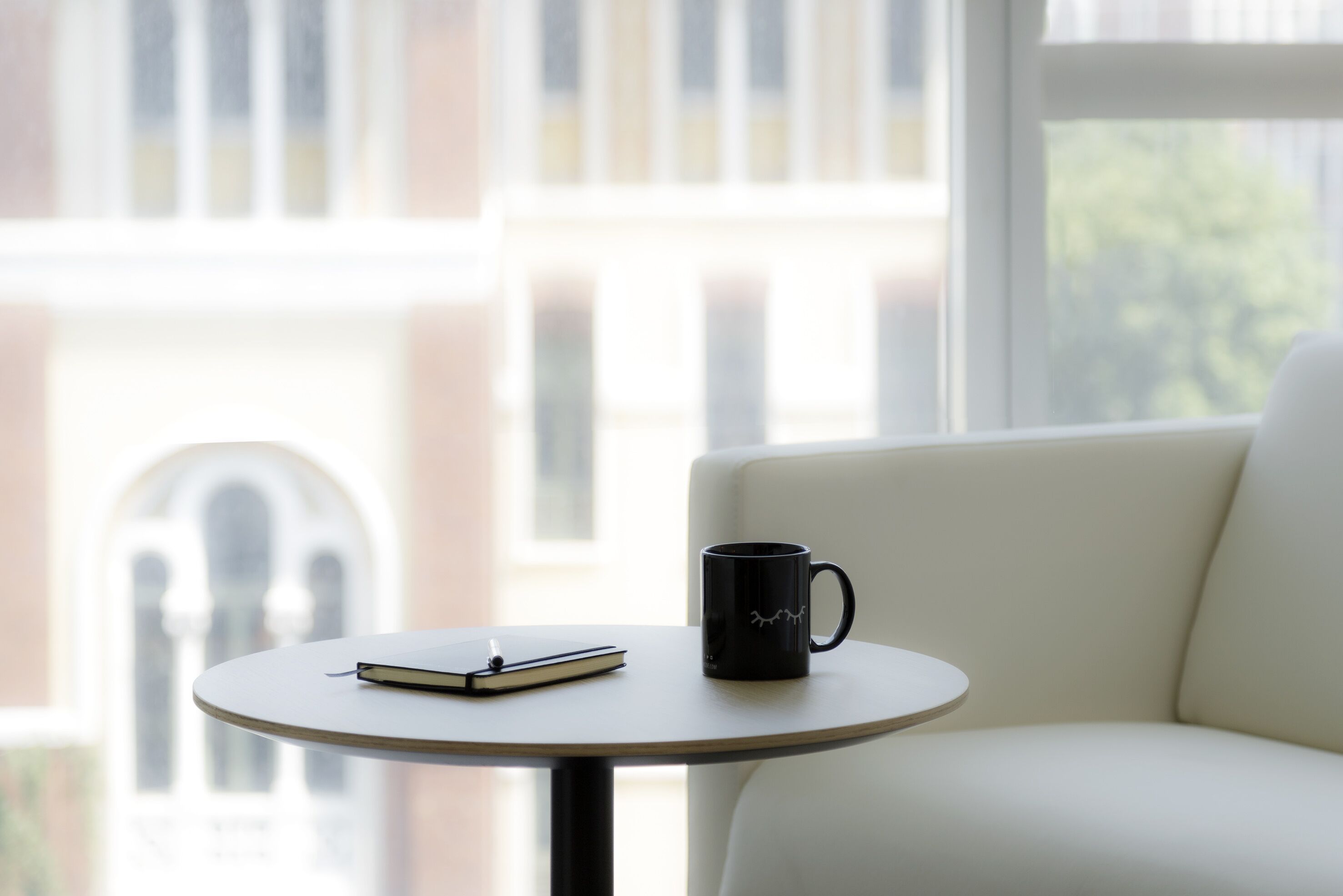 Una mesa redonda con una taza negra y un cuaderno, ubicada frente a una ventana que da a un edificio.