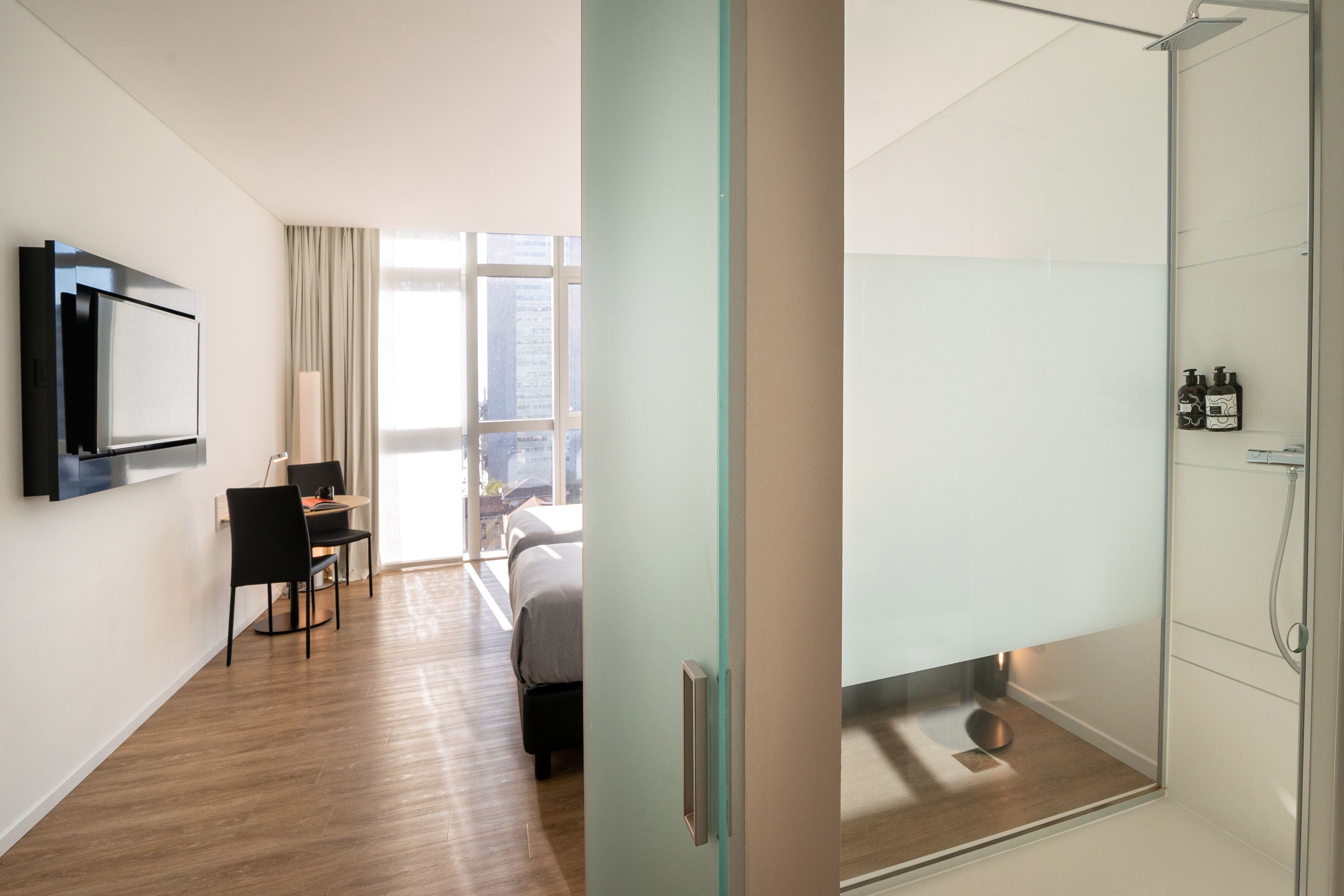 A modern hotel room with a wooden floor, a bed, a TV mounted on the wall, a dining area with a table and chairs, and a shower area with toiletries.