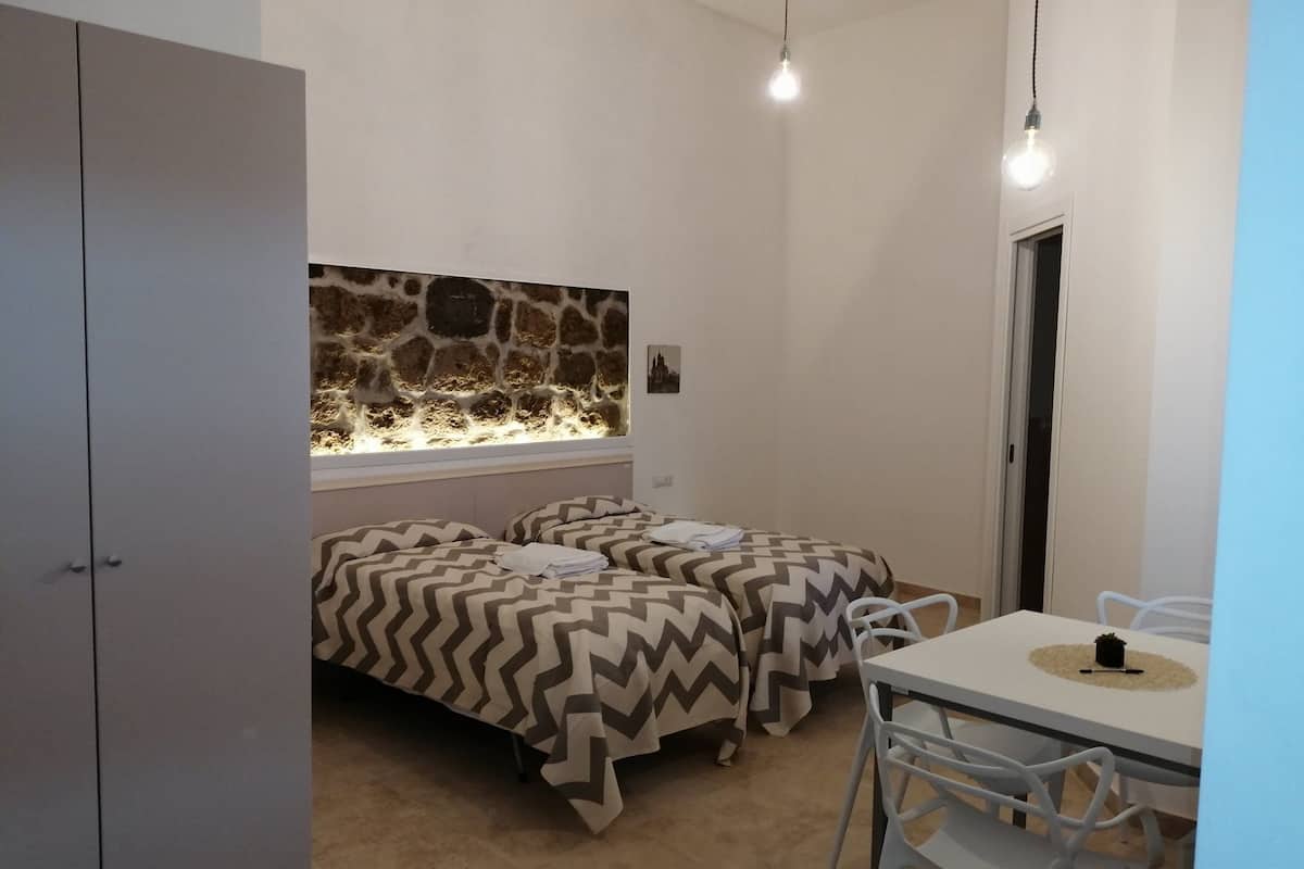 A small, well-lit room with two beds, a stone wall, a window, and a dining area with a table and chairs.