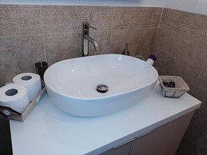 Un baño con un lavamanos blanco, un dispensador de papel higiénico y un pequeño recipiente con artículos de aseo.