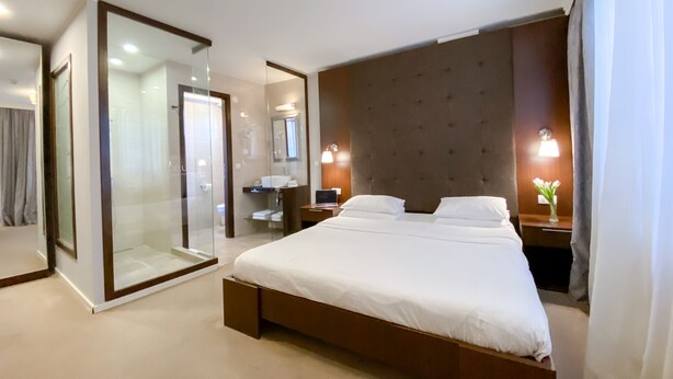 Een moderne hotelkamer met een groot bed, een douchecabine van glas, een kaptafel met spiegel en een nachtkastje met lamp.