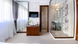 Hotelkamer met een grote spiegel, een flatscreen televisie, een houten bureau met een bloemenvaas en een douchecabine met glazen wanden.