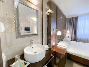 Een moderne hotelkamer met een grote ronde witte wastafel, een spiegel met decoratieve lijst, een nachtkastje met lamp en een keurig opgemaakt bed.